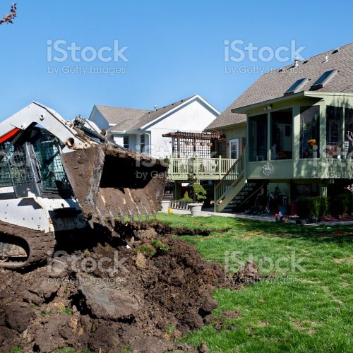 A small digger drops dirt removed from an excavation in back of a suburban house.